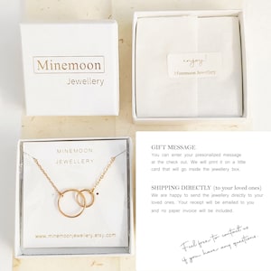 K&ouml;nnte beinhalten: Eine goldene Halskette mit zwei ineinander verschlungenen Kreisen, pr&auml;sentiert in einer wei&szlig;en Box mit dem Text "Minemoon Jewellery". Die Box befindet sich auf einer hellbraunen Oberfl&auml;che. Der Text "Z&ouml;gern Sie nicht, uns zu kontaktieren, wenn Sie Fragen haben." ist in schwarzer Tinte auf wei&szlig;em Hintergrund geschrieben.