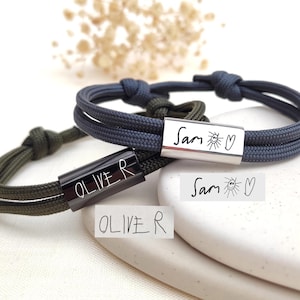 May include: Two personalised bracelets. One is navy blue with a silver bar engraved with "Sam" and a sun and heart symbol. The other is olive green with a black bar engraved with "Oliver". Both have a knotted design.