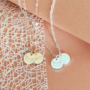 May include: Three gold and silver necklaces with angel wing charms and personalised name tags. The necklaces are on a peach-coloured fabric background.