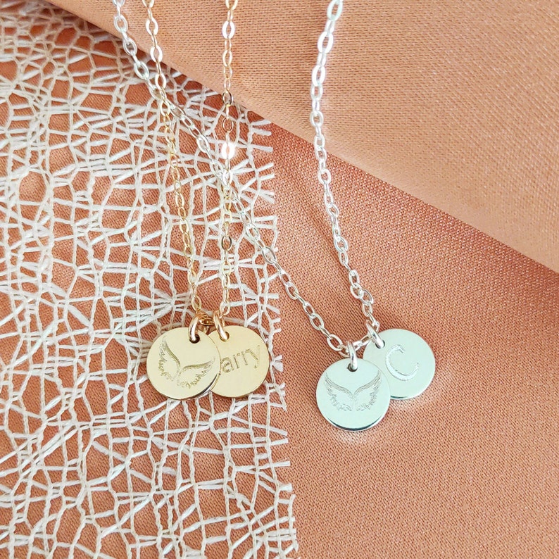 May include: Three gold and silver necklaces with angel wing charms and personalised name tags. The necklaces are on a peach-coloured fabric background.