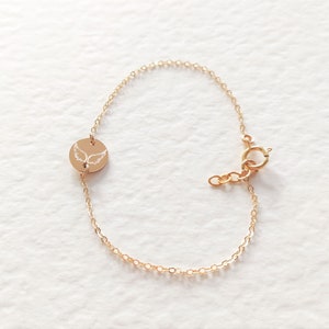 K&ouml;nnte beinhalten: Ein goldenes Kettenarmband mit einem kleinen runden Anh&auml;nger, der mit einem gefl&uuml;gelten Engelmotiv verziert ist.