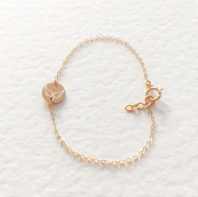 K&ouml;nnte beinhalten: Ein goldenes Kettenarmband mit einem kleinen runden Anh&auml;nger, der mit einem gefl&uuml;gelten Engelmotiv verziert ist.