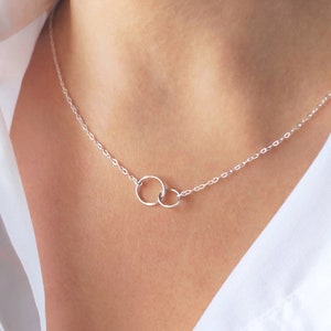 Könnte beinhalten: Eine zarte Silberkette mit zwei ineinandergreifenden Kreisen. Die Kreise sind durch eine feine Kette verbunden, wodurch ein minimalistisches und elegantes Design entsteht. Die Kette wird vor weißem Hintergrund gezeigt.