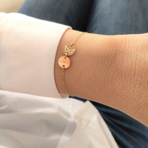 K&ouml;nnte beinhalten: Goldarmband mit zwei runden Anh&auml;ngern. Ein Anh&auml;nger hat ein Datum eingraviert, der andere Anh&auml;nger hat ein Paar Engelsfl&uuml;gel eingraviert.