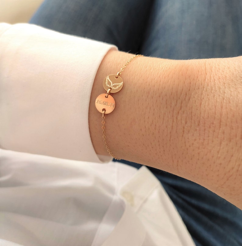 K&ouml;nnte beinhalten: Goldarmband mit zwei runden Anh&auml;ngern. Ein Anh&auml;nger hat ein Datum eingraviert, der andere Anh&auml;nger hat ein Paar Engelsfl&uuml;gel eingraviert.