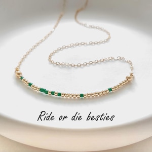 May include: A gold-tone necklace with a curved bar pendant. The pendant is adorned with small gold beads and green square beads. The necklace is presented on a white surface. The image includes the text "Ride or die besties."