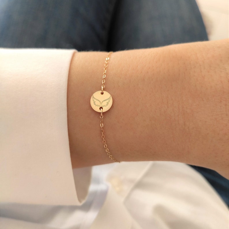 K&ouml;nnte beinhalten: Ein goldenes Kettenarmband mit einem kleinen runden Anh&auml;nger, der mit einem gravierten Engelsfl&uuml;gelmotiv verziert ist.