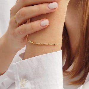 May include: A delicate gold bracelet with small beads and a chain. The bracelet is worn on a person's wrist.