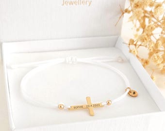 May include: A white cord bracelet with a gold cross pendant, two gold beads, and a small gold heart charm. The cross is engraved with the name "Sophie" and the date "23.4.2023". The bracelet is presented in a white box.