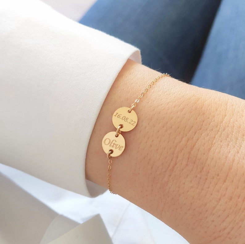 K&ouml;nnte beinhalten: Ein goldenes Kettenarmband mit zwei runden Anh&auml;ngern. Ein Anh&auml;nger ist mit dem Datum "16.08.22" graviert und der andere Anh&auml;nger ist mit dem Namen "Olive" graviert.