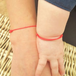 Könnte beinhalten: Zwei Handgelenke mit roten Schnurarmbändern. Die Armbänder sind mit einem einfachen Knoten gebunden. Die Handgelenke liegen nebeneinander, eines ist ein Erwachsenenhandgelenk und das andere ein Kinderhandgelenk.