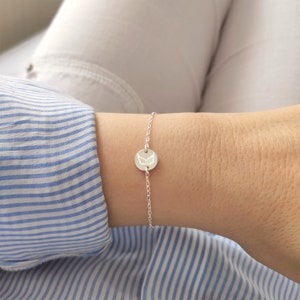 K&ouml;nnte beinhalten: Ein silbernes Kettenarmband mit einem kleinen runden Anh&auml;nger mit einem gravierten Engelsfl&uuml;gel-Design.