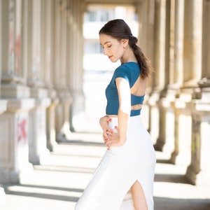 Könnte beinhalten: Eine Frau trägt ein blaues Crop-Top mit einer weißen Rock und schwarzen High Heels. Der Rock hat einen Schlitz an der Seite. Sie steht in einem Flur mit weißen Säulen.