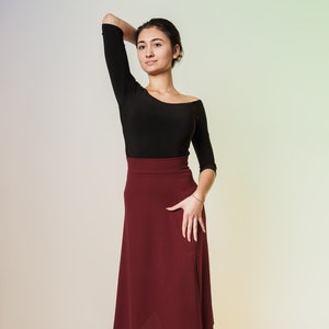 Könnte beinhalten: Eine Frau trägt ein schwarzes Langarmshirt und einen burgunderfarbenen Midirock. Sie steht mit der Hand auf der Hüfte und schaut zur Seite.
