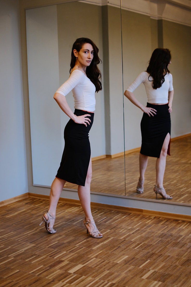 K&ouml;nnte beinhalten: Eine Frau in einem wei&szlig;en Oberteil und einem schwarzen Rock mit seitlichem Schlitz steht vor einem Spiegel. Sie tr&auml;gt nude High Heels und hat ihre Haare nach hinten gebunden.