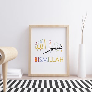 Op de afbeelding: Een ingelijst afdruk met de Arabische zin "Bismillah" in kleurrijke kalligrafie. De zin is in zwart geschreven met kleurrijke accenten. De lijst is van licht hout en de achtergrond is wit.