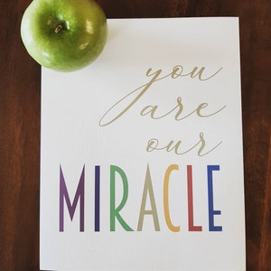 May include: A white card with gold lettering that says "you are our MIRACLE" in rainbow colors. A green apple is in the top left corner of the image.