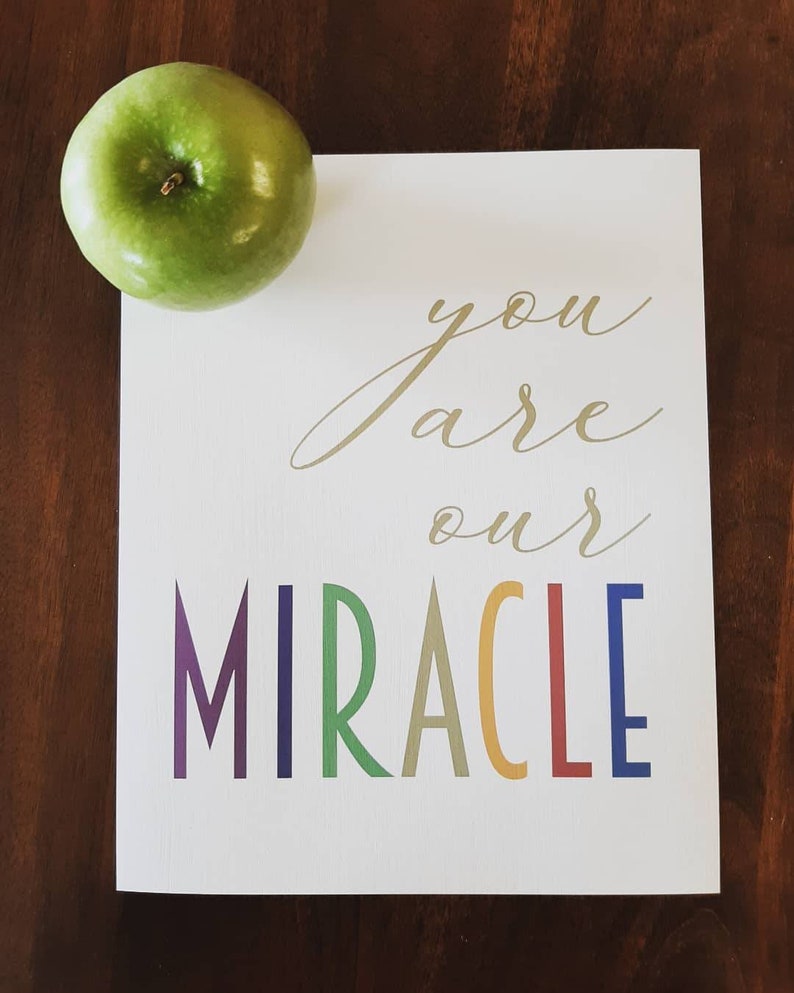 May include: A white card with gold lettering that says "you are our MIRACLE" in rainbow colors. A green apple is in the top left corner of the image.