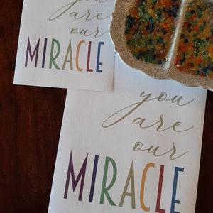 May include: Two white cards with the text "you are our MIRACLE" in gold script and rainbow colored letters. The cards are on a wooden surface.
