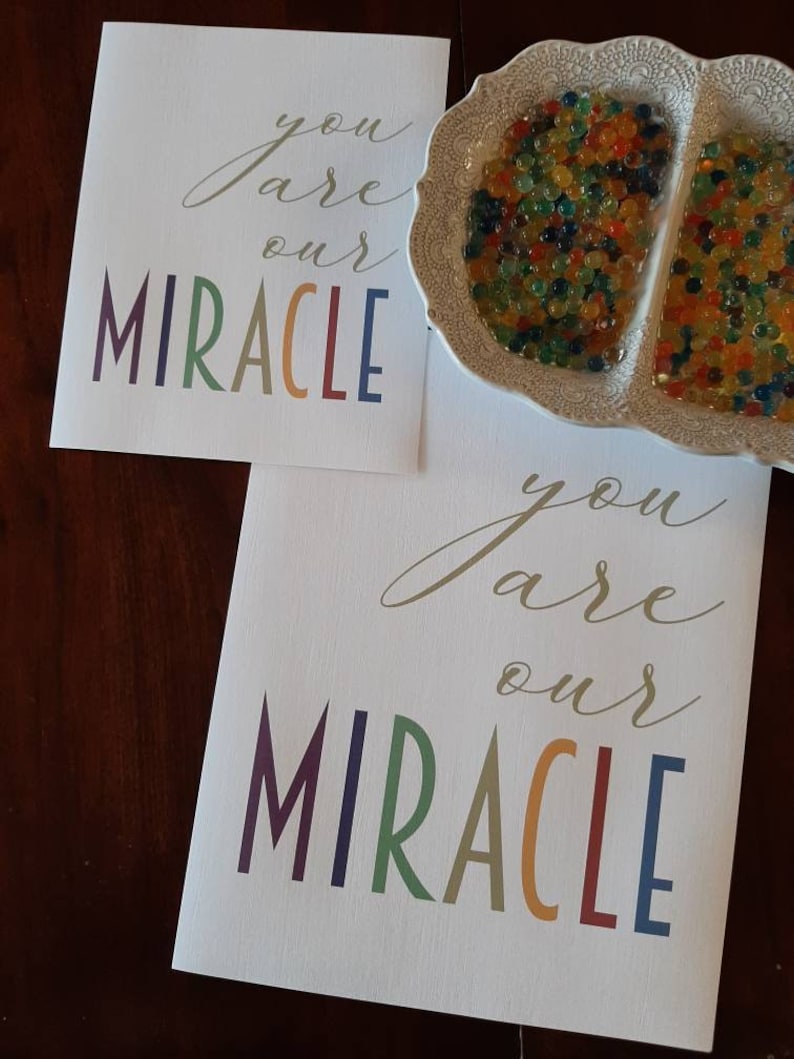 May include: Two white cards with the text "you are our MIRACLE" in gold script and rainbow colored letters. The cards are on a wooden surface.