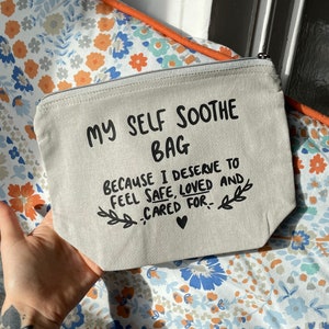 May include: A beige canvas zippered pouch featuring the black printed text MY SELF SOOTHE BAG, BECAUSE I DESERVE TO FEEL SAFE, LOVED AND CARED FOR, accompanied by two small leafy branch illustrations and a tiny heart symbol centred at the bottom.