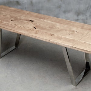 May include: A long, rectangular wooden table with a natural wood finish and a live edge. The table has a metal frame with a polished chrome finish. The table is sitting on a concrete floor.