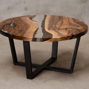 May include: Round wooden coffee table with a black metal base. The table top has a natural wood grain pattern and a clear epoxy resin fill with a silver and black swirl.