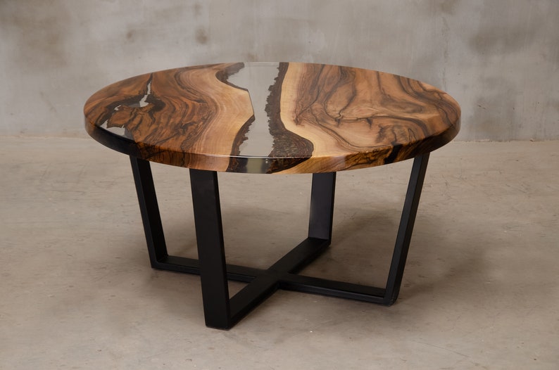 May include: Round wooden coffee table with a black metal base. The table top has a natural wood grain pattern and a clear epoxy resin fill with a silver and black swirl.