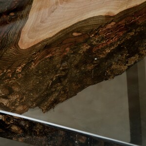 K&ouml;nnte beinhalten: Nahaufnahme einer Tischplatte aus Holz mit lebendiger Kante. Die Holzmaserung weist eine Mischung aus hellbraunen, dunkelbraunen und schwarzen T&ouml;nen auf. Die Tischplatte steht auf einer transparenten Glasoberfl&auml;che mit einem verchromten Metallrahmen.