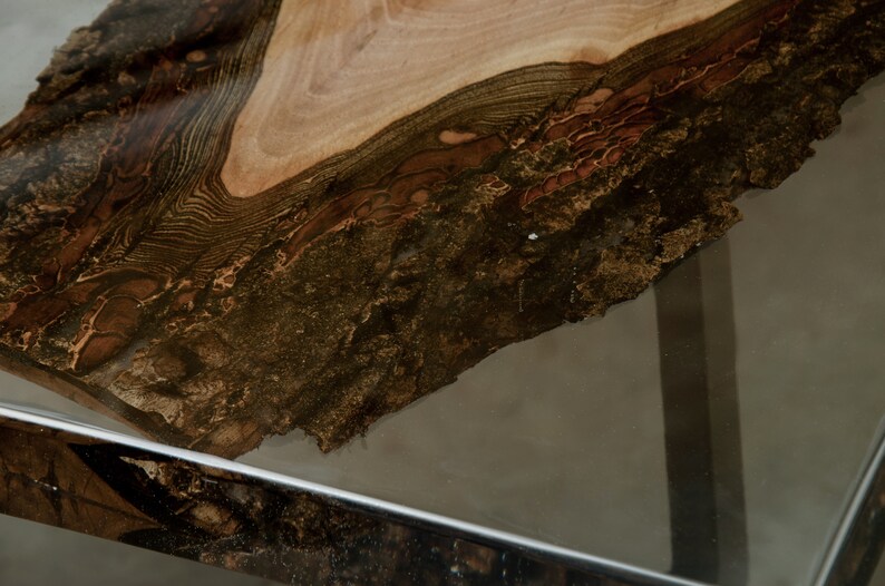 K&ouml;nnte beinhalten: Nahaufnahme einer Tischplatte aus Holz mit lebendiger Kante. Die Holzmaserung weist eine Mischung aus hellbraunen, dunkelbraunen und schwarzen T&ouml;nen auf. Die Tischplatte steht auf einer transparenten Glasoberfl&auml;che mit einem verchromten Metallrahmen.