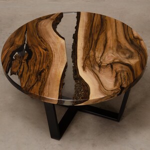 May include: Round wooden coffee table with a clear epoxy resin inlay. The table has a black metal base with a cross-shaped design.