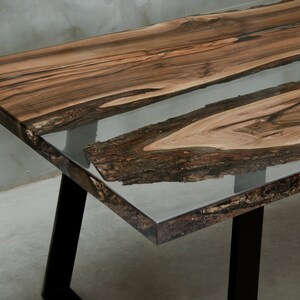 K&ouml;nnte beinhalten: Handgefertigter Holztisch mit einem Design mit lebendiger Kante. Der Tisch hat einen transparenten Epoxidharzfluss in der Mitte und schwarze Metallbeine. Das Holz hat eine nat&uuml;rliche, dunkelbraune Farbe. Ca. 150 cm lang.