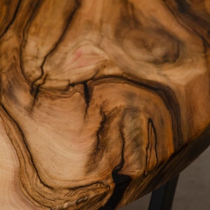May include: Close-up of a round wooden table top with a natural wood grain pattern. The wood is a rich brown color with dark brown streaks. The table top is smooth and polished.