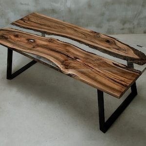 K&ouml;nnte beinhalten: Ein rechteckiger Couchtisch aus Holz mit einer transparenten Harzmitte. Der Tisch hat ein nat&uuml;rliches Holzmaserungsmuster in Braun- und Schwarzt&ouml;nen. Er hat eine klare, gl&auml;nzende Oberfl&auml;che und steht auf schwarzen Metallbeinen. Der Tisch ist etwa 152 cm lang.
