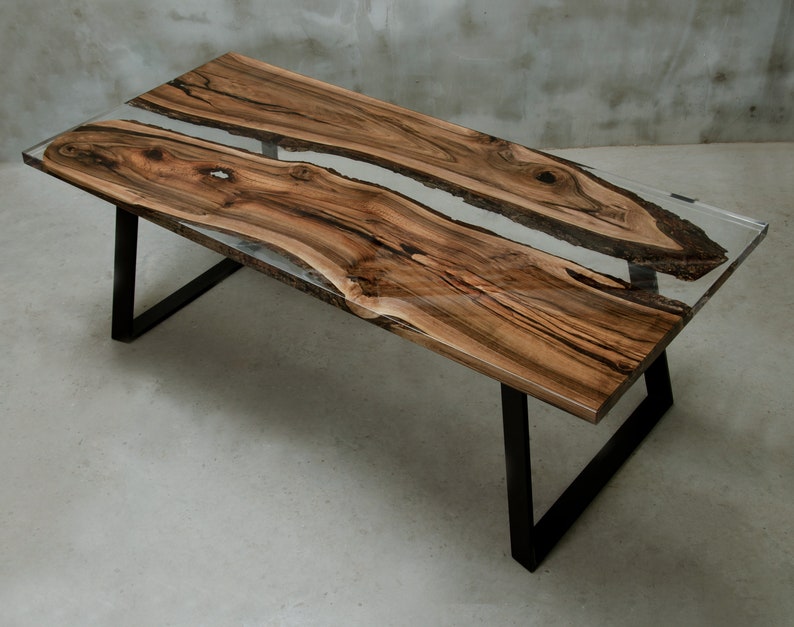 K&ouml;nnte beinhalten: Ein rechteckiger Couchtisch aus Holz mit einer transparenten Harzmitte. Der Tisch hat ein nat&uuml;rliches Holzmaserungsmuster in Braun- und Schwarzt&ouml;nen. Er hat eine klare, gl&auml;nzende Oberfl&auml;che und steht auf schwarzen Metallbeinen. Der Tisch ist etwa 152 cm lang.