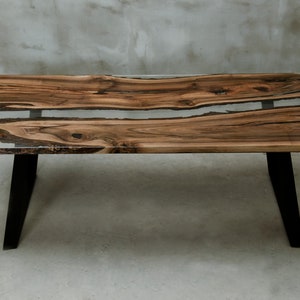 K&ouml;nnte beinhalten: Ein rechteckiger Couchtisch mit einer Tischplatte aus Holz mit lebendiger Kante und einem Inlay aus klarem Harz. Der Tisch hat schwarze Metallbeine. Das Holz hat ein nat&uuml;rliches Maserungsmuster mit Braun- und Beiget&ouml;nen. Der Tisch ist etwa 122 cm lang.