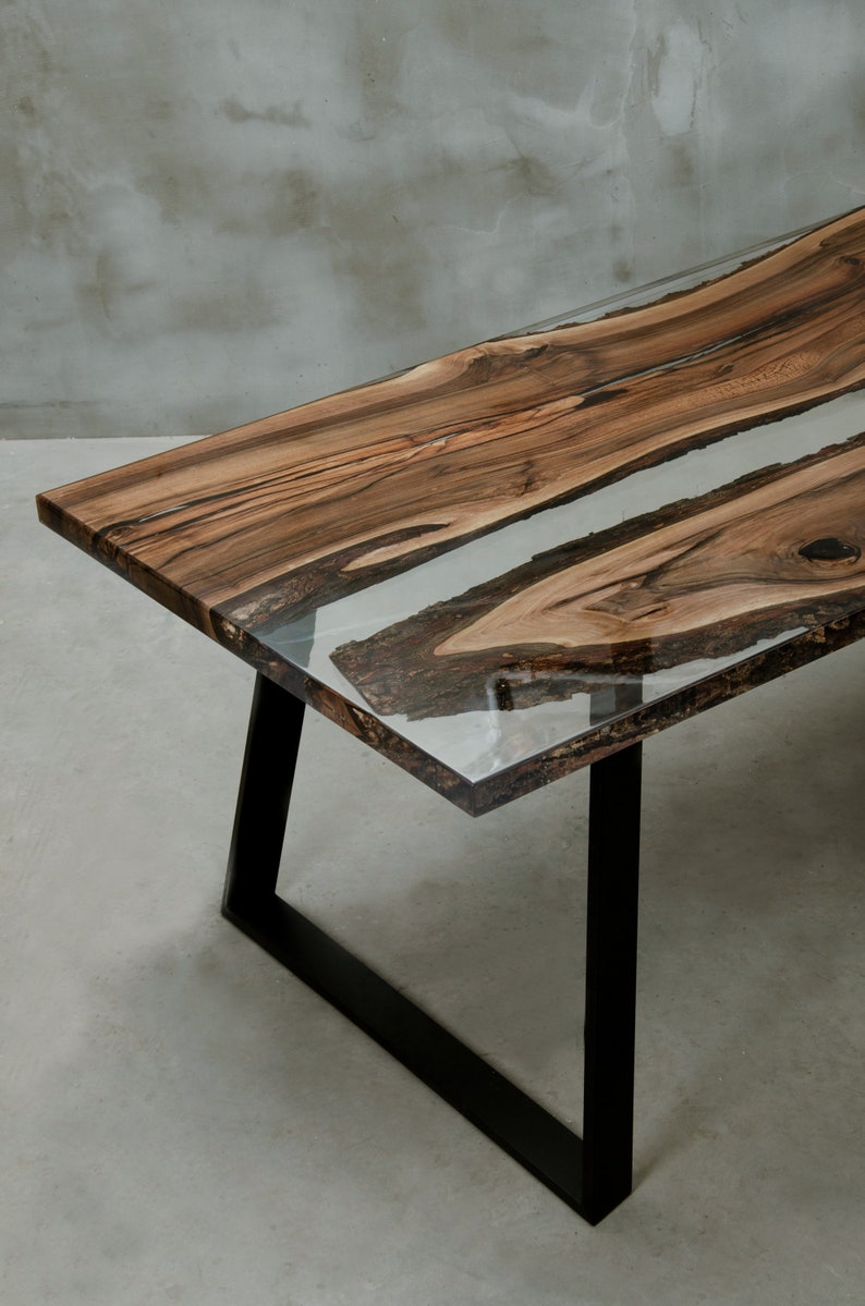 K&ouml;nnte beinhalten: Ein handgefertigter Holztisch mit einem Design mit lebendiger Kante. Die Tischplatte verf&uuml;gt &uuml;ber einen transparenten Epoxidharzfluss, der durch die Mitte verl&auml;uft. Der Tisch wird von schwarzen Metallbeinen getragen. Abmessungen: ca. 183 cm lang, 91 cm breit und 76 cm hoch.