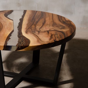 May include: A round wooden coffee table with a natural wood grain finish and a clear epoxy resin inlay. The table has a black metal base with a geometric design.