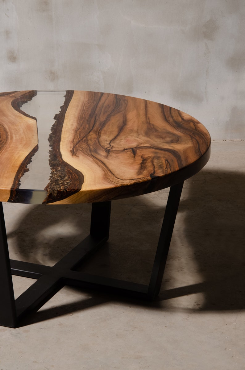 May include: A round wooden coffee table with a natural wood grain finish and a clear epoxy resin inlay. The table has a black metal base with a geometric design.