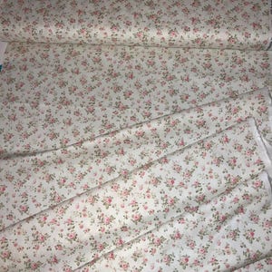 May include: A roll of fabric with a repeating pattern of small pink roses and green leaves on a cream background. The fabric is folded and rolled, showing the floral design in detail. Ideal for sewing projects.