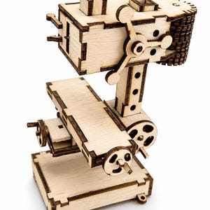 Op de afbeelding: Een houten model van een machine met ingewikkelde details. Het model heeft een rechthoekige basis, een centrale kolom met een groot wiel en een bovendeel met een tandwielachtig ontwerp. Het hout is licht van kleur met donkere randen.