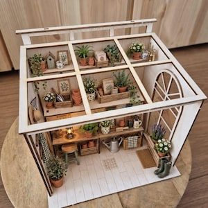Op de afbeelding: Een miniatuur houten kasmodel met een wit frame, tegelvloer en planken vol kleine potplanten, tuingereedschap, zaadzakjes, een klein krukje, een metalen gieter, een lantaarn en een paar groene rubberen laarzen.