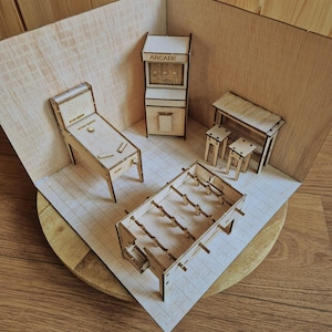 Op de afbeelding: Een miniatuur houten speelhalset met een flipperkast met het label Star Quest, een staande arcadekast, een tafelvoetbalspel en een statafel met twee krukken. De set is gemaakt van lichtgekleurd multiplex met lasergesneden details.
