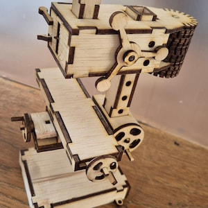 Puede incluir: Maqueta de madera de una máquina compleja. Presenta una base rectangular, un soporte vertical y una sección superior elaborada con engranajes, palancas y una pequeña chimenea. La madera tiene un acabado natural con bordes más oscuros.