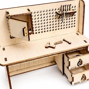 May include: A miniature wooden workbench with a pegboard back, a cabinet with an open door, and two drawers. The workbench has small wooden tools on the surface, including a hammer, wrench, and screwdriver. The wood is light-coloured.
