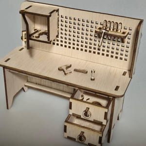 Könnte beinhalten: Eine Miniatur-Werkbank aus hellem Holz mit Lochwand, Schubladen und kleinen Werkzeugen. Die Werkbank besteht aus hellem Holz mit dunkleren Kanten. Die Lochwand hat gleichmäßig verteilte Löcher. Zu den Werkzeugen gehören Hammer, Schraubenschlüssel und andere kleine Gegenstände.