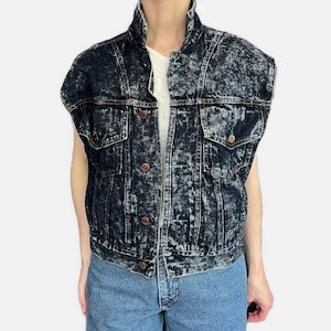 Puede incluir: Chaleco vaquero azul oscuro con lavado ácido, cuello alto y cierre de botones. El chaleco tiene dos bolsillos en el pecho y dos bolsillos laterales. El denim tiene un patrón moteado. Los botones son de color cobre.