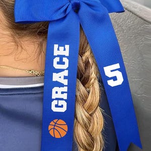 May include: A royal blue hair bow with the name "GRACE" in white block letters and an orange basketball graphic. The bow also features the number "5" in white. The bow is tied in a knot and is worn in a braid.