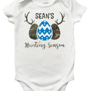 May include: White baby onesie with a blue and white patterned Easter egg, two camouflage eggs, and antlers. The text "SEAN'S Hunting Season" is printed on the onesie.