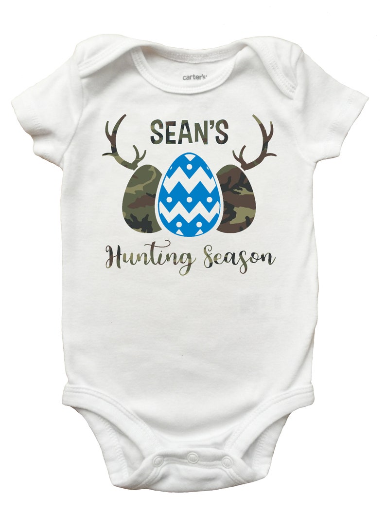 May include: White baby onesie with a blue and white patterned Easter egg, two camouflage eggs, and antlers. The text "SEAN'S Hunting Season" is printed on the onesie.